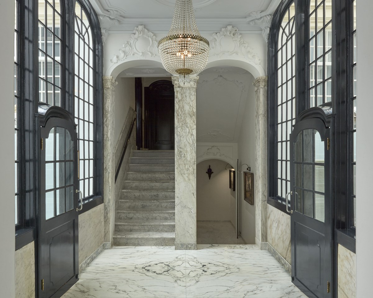 Grand marble hallway with a chandelier, tall arched windows, and stairs leading down at Pulitzer Amsterdam.