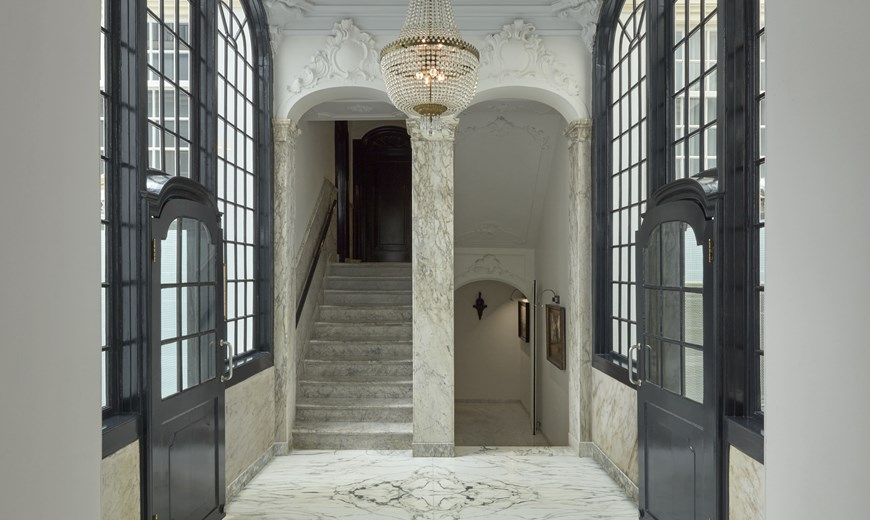 Grand marble hallway with a chandelier, tall arched windows, and stairs leading down at Pulitzer Amsterdam.