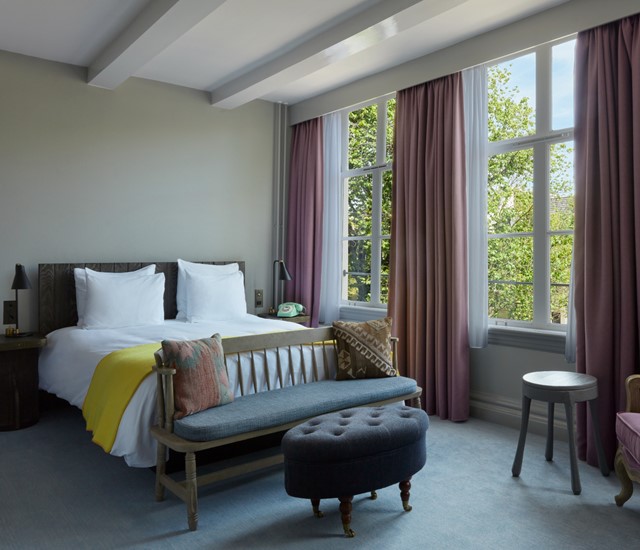 A cozy hotel room featuring a bed, a chair, a table, and a window allowing natural light to enter the space.