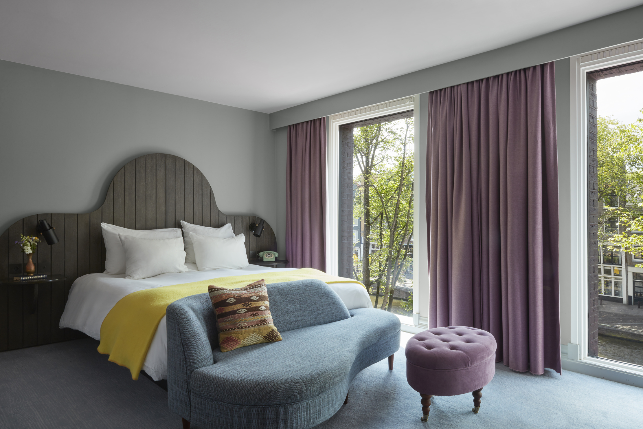 Elegant room with a curved wooden headboard, blue sofa, and purple curtains overlooking trees and a canal.