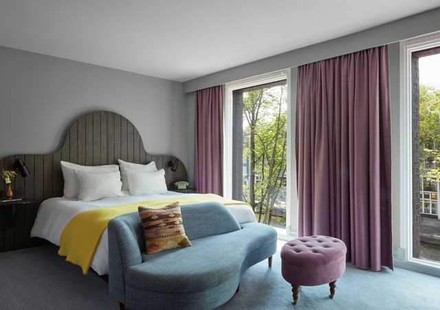 Elegant room with a curved wooden headboard, blue sofa, and purple curtains overlooking trees and a canal.