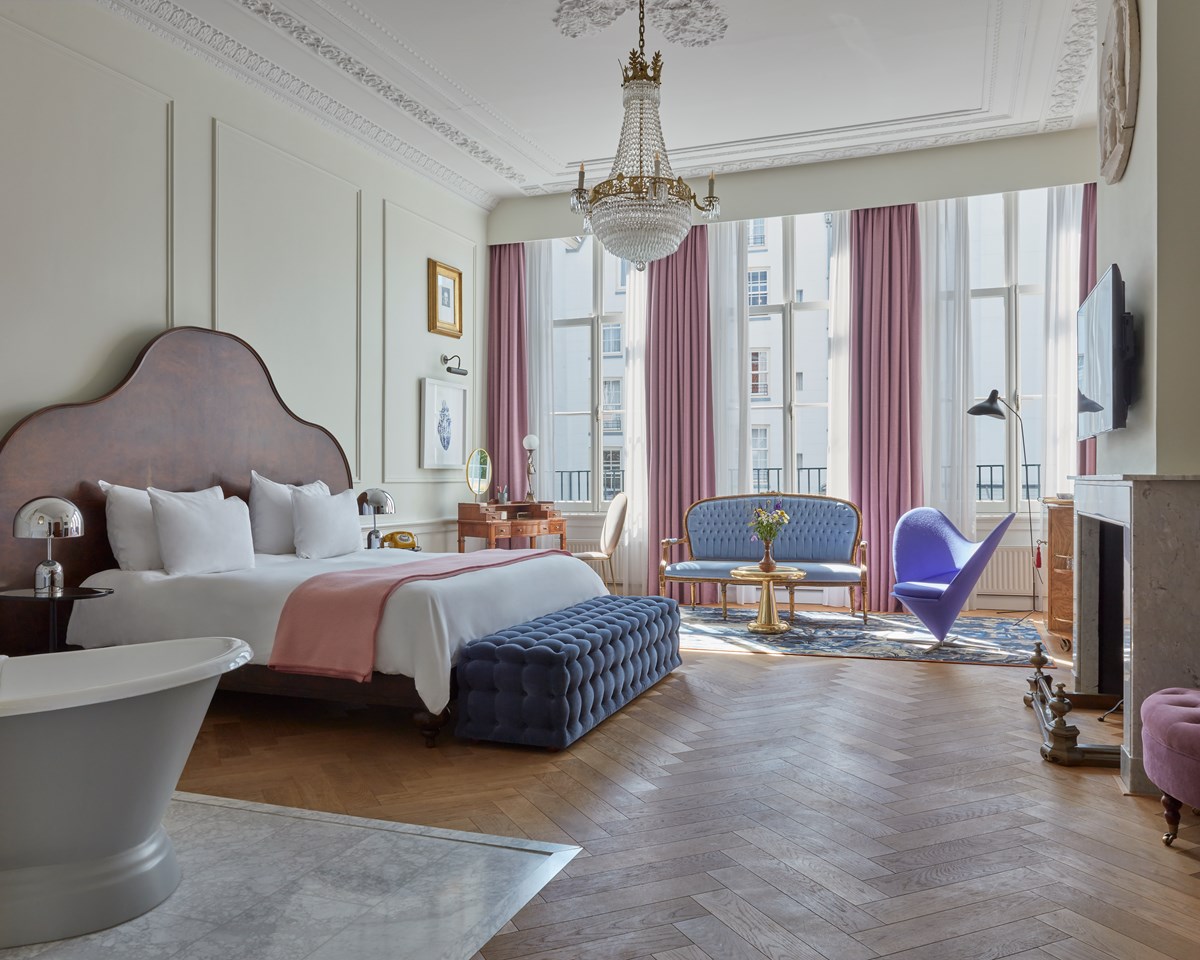 Elegant Pulitzer Amsterdam hotel suite with a freestanding bathtub, large bed, chandelier, tall windows and pink curtains.
