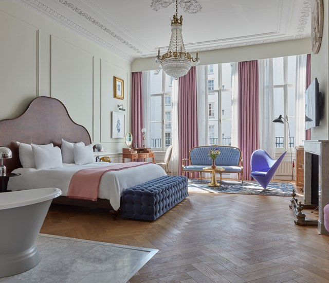 Elegant Pulitzer Amsterdam hotel suite with a freestanding bathtub, large bed, chandelier, tall windows and pink curtains.