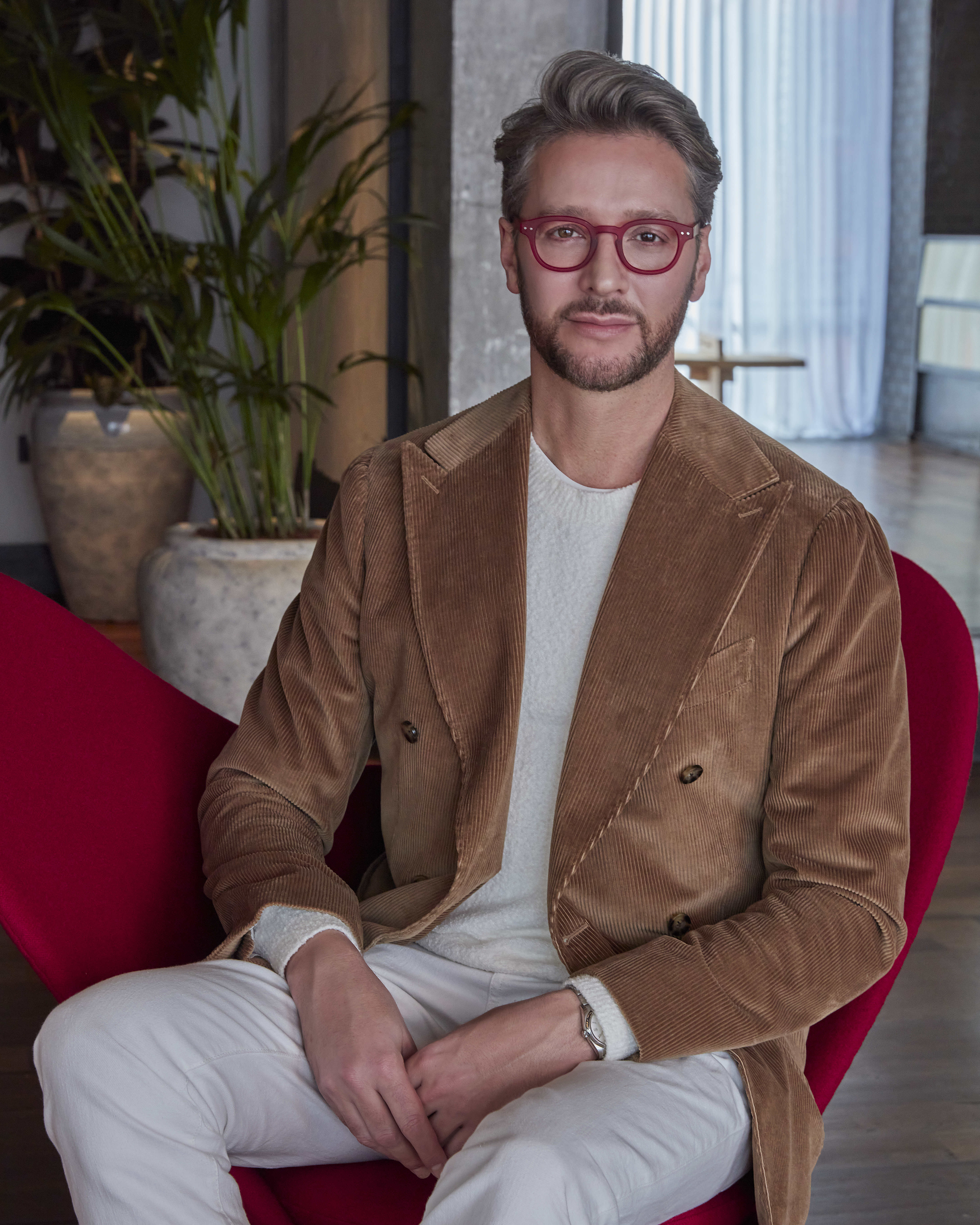 Jacu Strauss seated in a modern chair wearing a brown blazer, white pants, and red glasses in a stylish hotel setting.