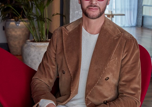 Jacu Strauss seated in a modern chair wearing a brown blazer, white pants, and red glasses in a stylish hotel setting.