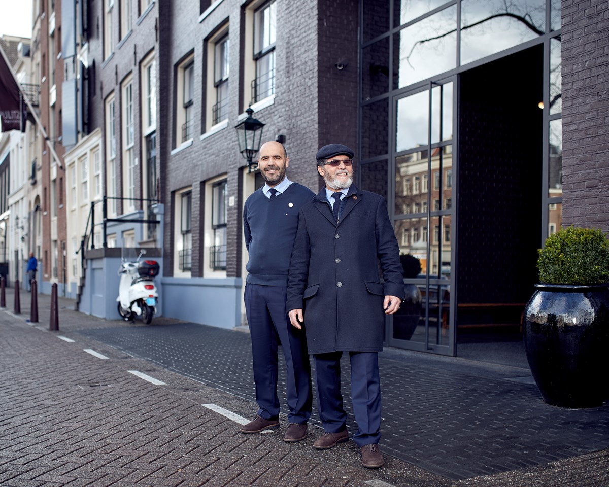 Two men positioned on a brick sidewalk, seemingly in dialogue, with a backdrop of a peaceful street scene.