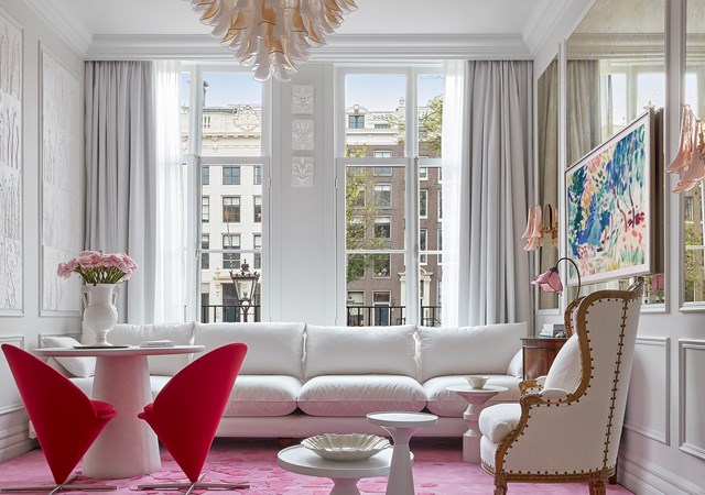 Stylish lounge with modern red chairs, white sofa, and large windows overlooking Amsterdam's historic architecture.