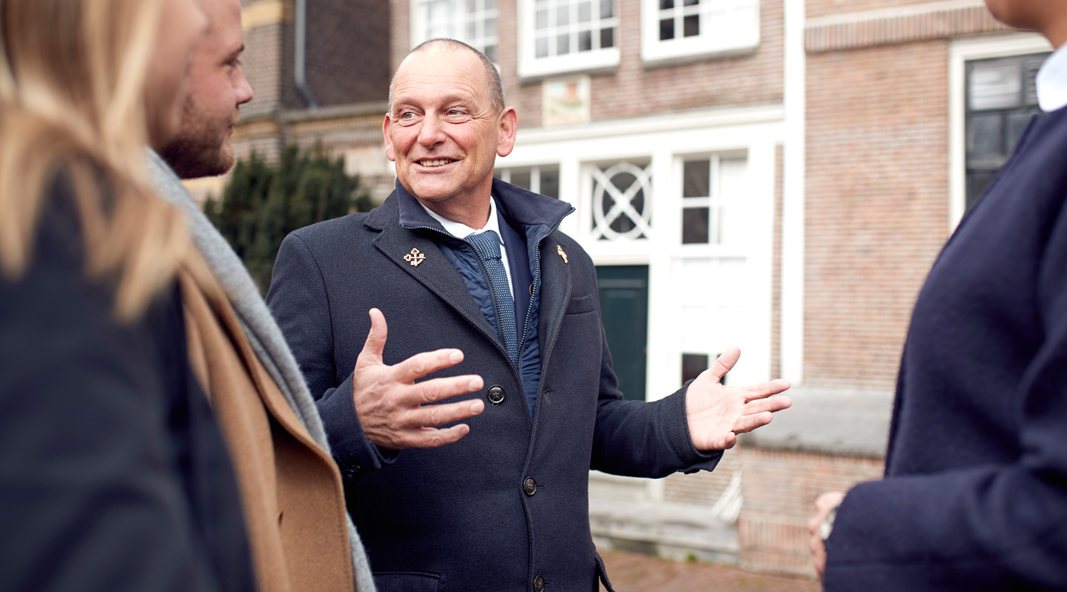 Hotel concierge in a navy coat with an emblem, speaking with hands gesturing outside of an Amsterdam building.