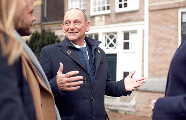 Hotel concierge in a navy coat with an emblem, speaking with hands gesturing outside of an Amsterdam building.