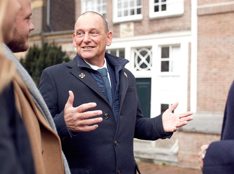 Hotel concierge in a navy coat with an emblem, speaking with hands gesturing outside of an Amsterdam building.