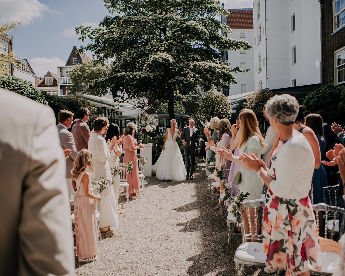 A wedding ceremony in a courtyard, with guests standing in the aisle, surrounded by greenery and elegant decorations.