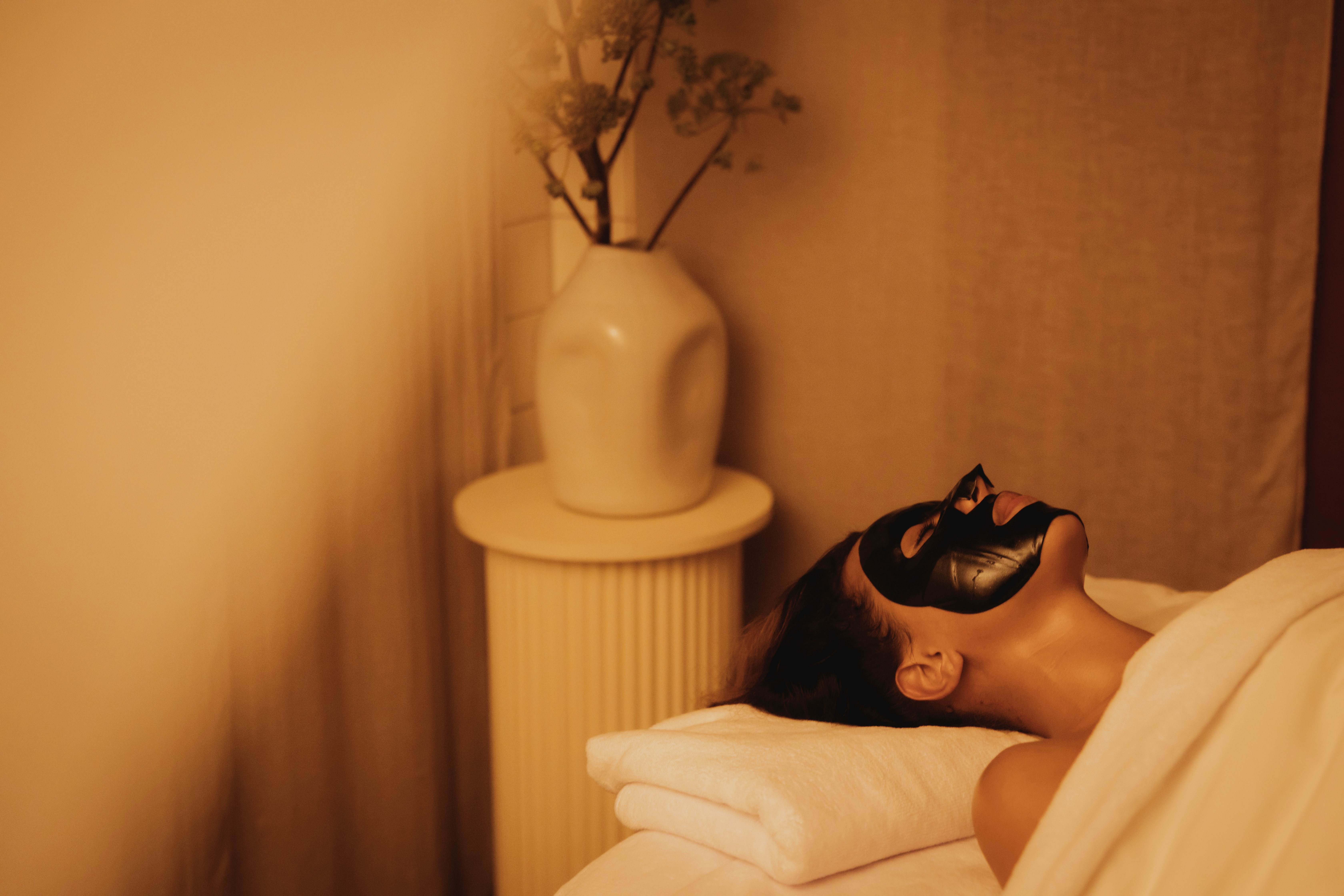 A woman wearing a black face mask, relaxing on a massage table in a peaceful and relaxing space. 