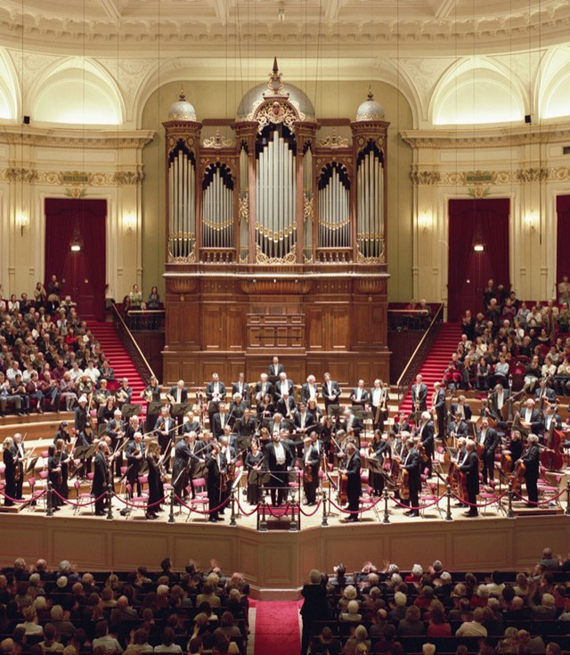 A large orchestra performs in a spacious hall, led by a conductor, creating a harmonious musical experience.