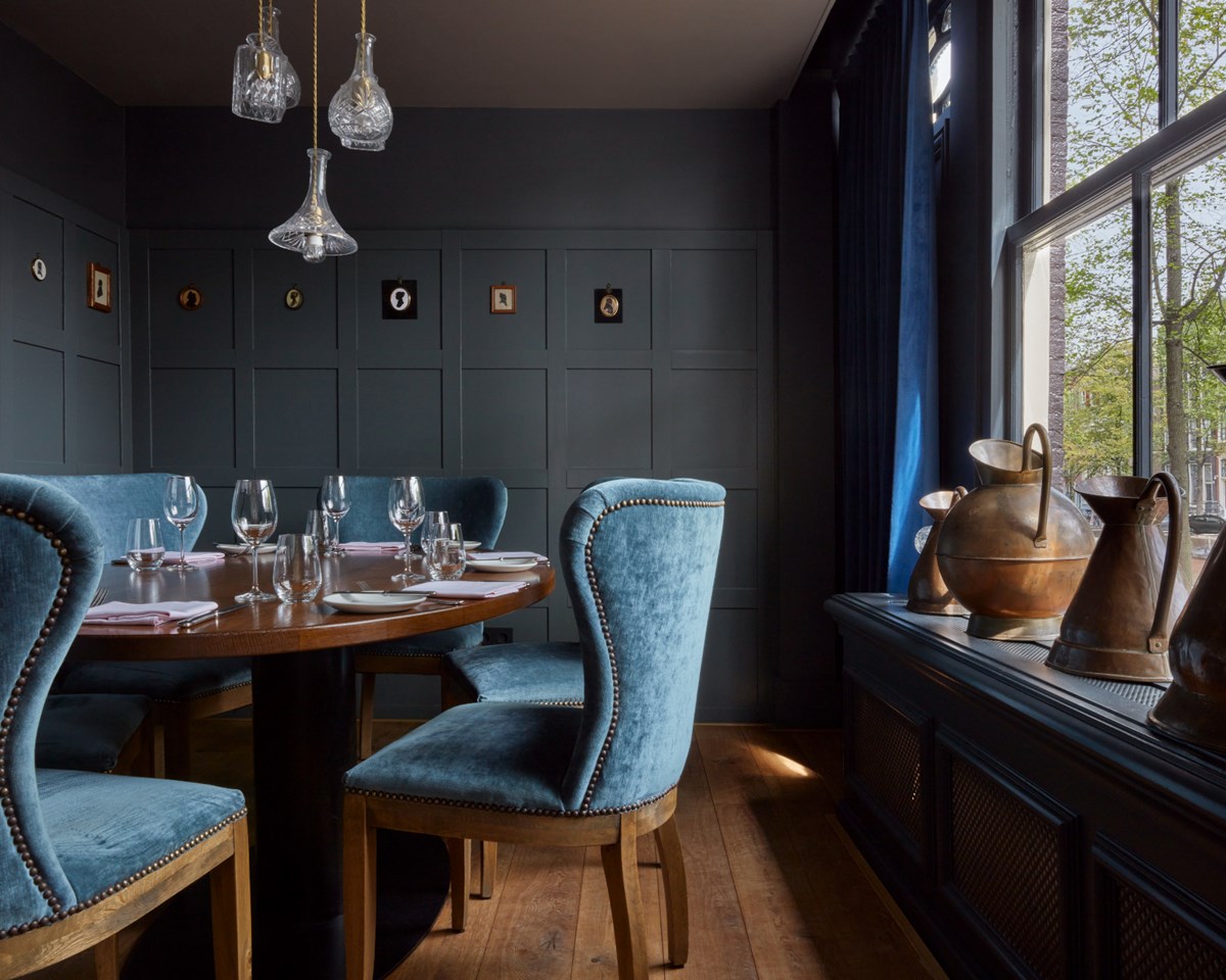 Intimate dining area with blue velvet chairs, a wooden table set with wine glasses, and brass vases by a window.