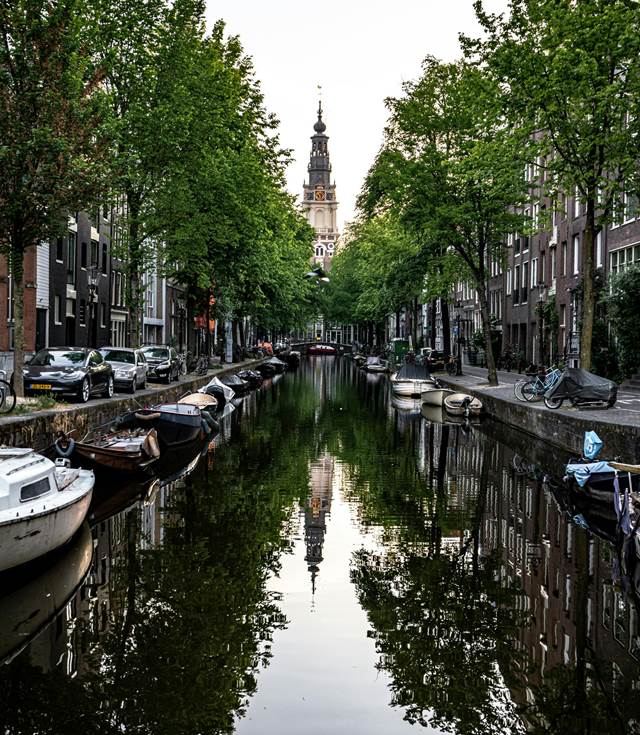 Amsterdam canal lined with trees and boats, with a historic church steeple in the background, reflected in the water.