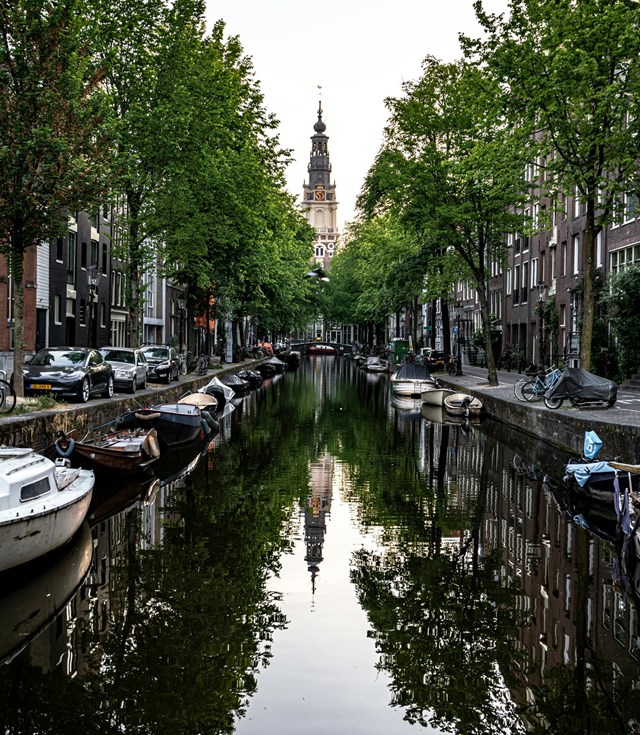 Amsterdam canal lined with trees and boats, with a historic church steeple in the background, reflected in the water.