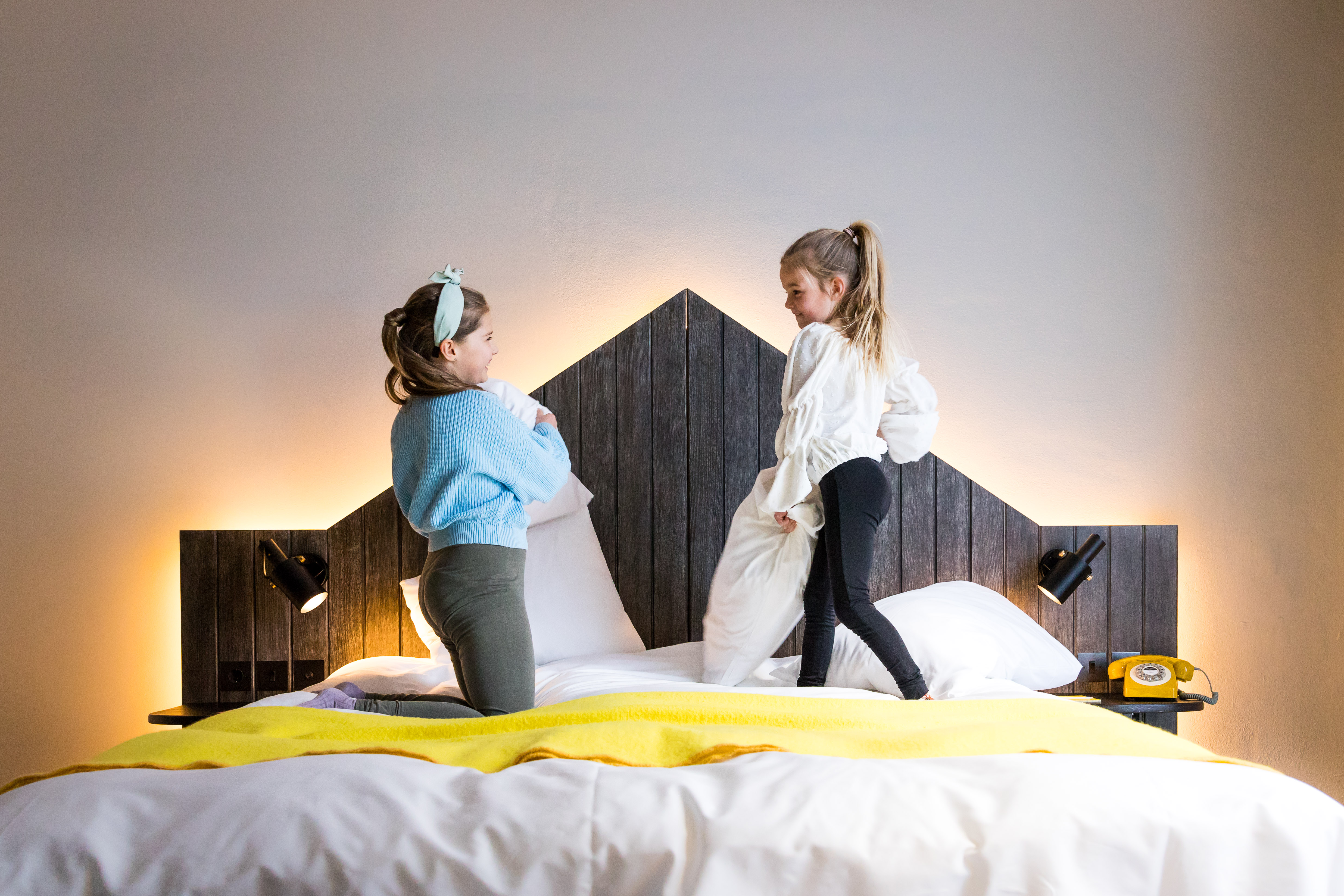 Two girls stand on a bed covered with a bright yellow blanket, smiling and enjoying their time together.