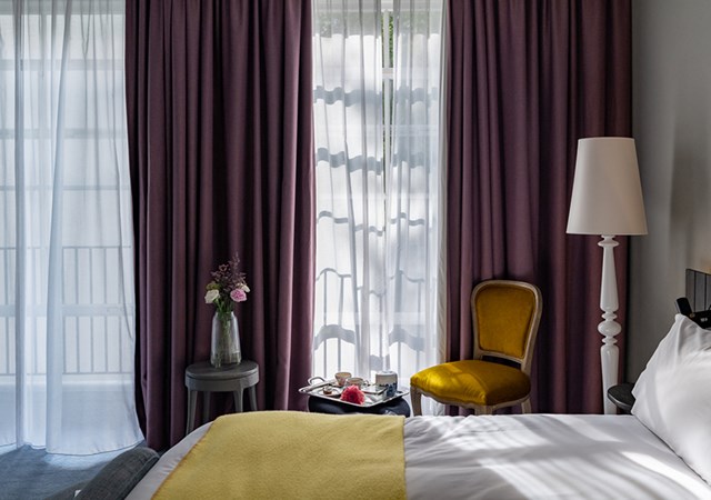 Cozy hotel bedroom with a yellow blanket, purple curtains, and natural light filtering through sheer drapes.