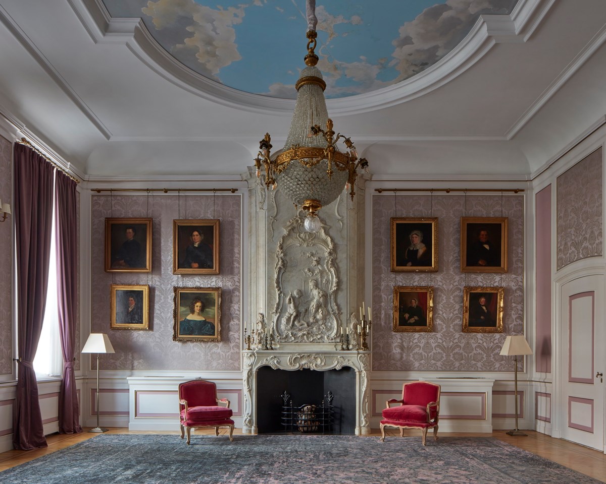 Elegant lounge with a chandelier, classical portraits, red velvet chairs, and a white fireplace under a painted sky ceiling.