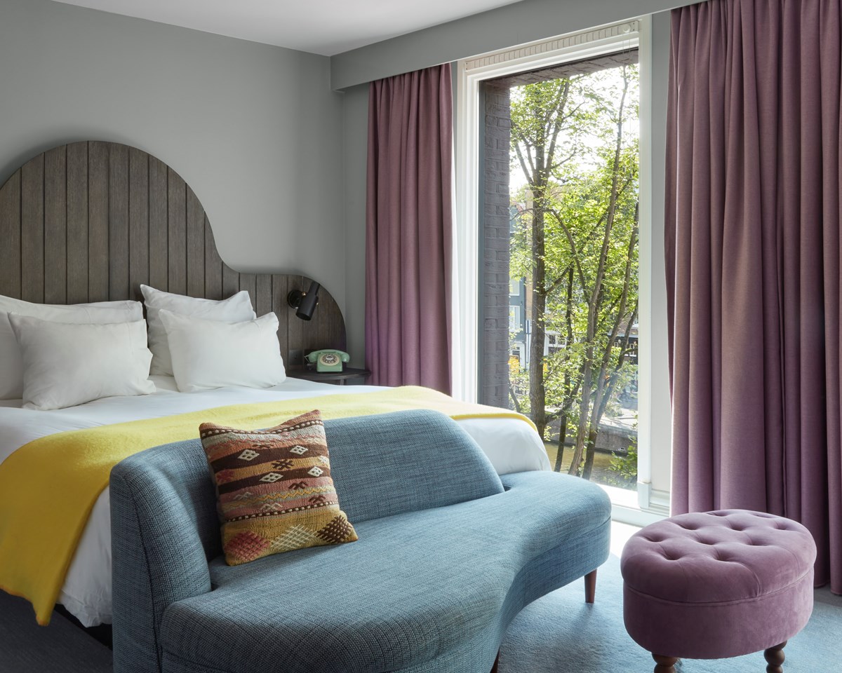 Pulitzer Amsterdam bedroom featuring a curved headboard, yellow throw blanket, and a cozy seating area by large windows.