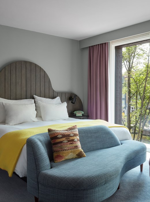 Pulitzer Amsterdam bedroom featuring a curved headboard, yellow throw blanket, and a cozy seating area by large windows.