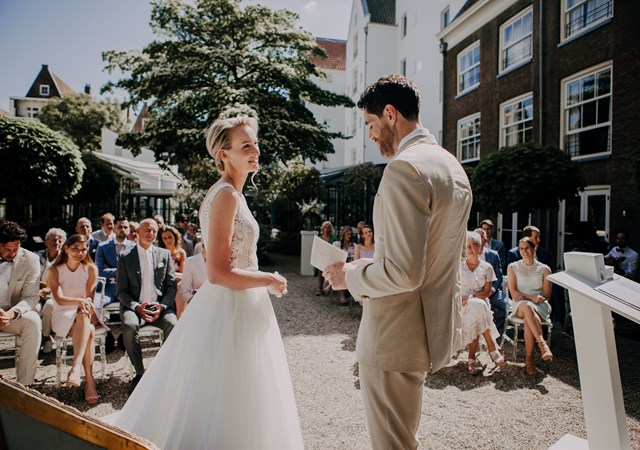 Bride and groom standing outdoors during their wedding ceremony, surrounded by guests and summer greenery.