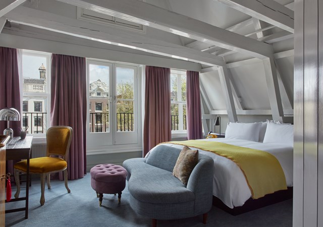 Cozy attic-style hotel room with a large bed, purple curtains, and windows overlooking classic Amsterdam architecture.