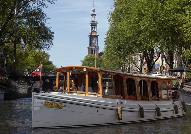 Classic canal boat, the Pulitzer Boat with wooden cabin cruising through Amsterdam with Westerkerk tower in the background.