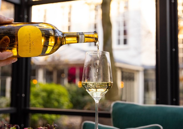 Hand pouring white wine into a glass on a table in a lush garden setting with green seating at Pulitzer's Garden.