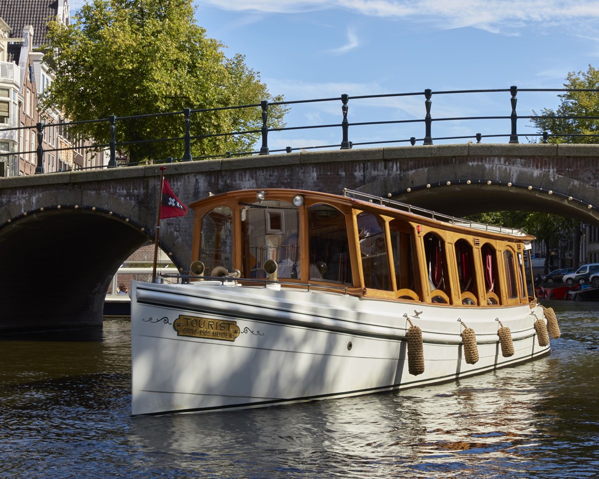 A scenic Amsterdam canal view with the Pulitzer Boat sailing under a bridge with a clear blue sky.
