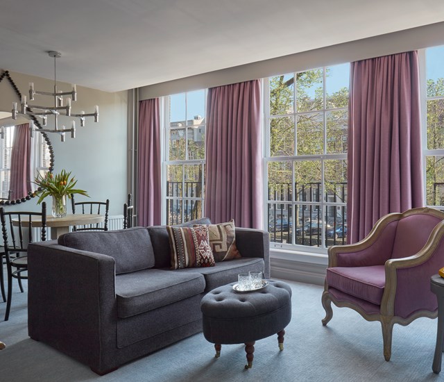 Stylish hotel suite at Pulitzer Amsterdam with gray sofa, mauve curtains, and a purple armchair by large sunny windows.