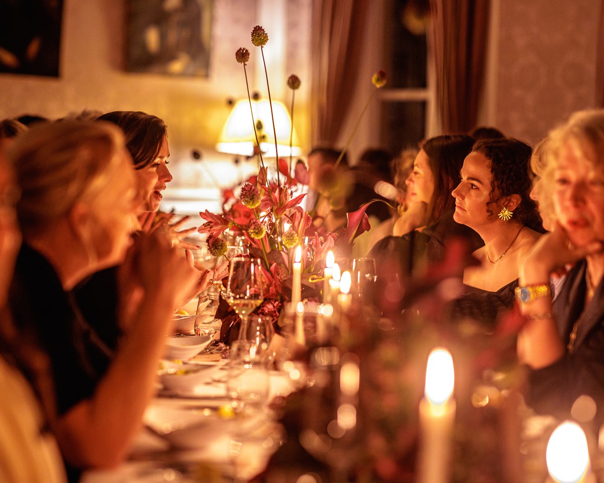 Women seated at a long, candle-lit dinner table, engaged in conversation and surrounded by floral arrangements.